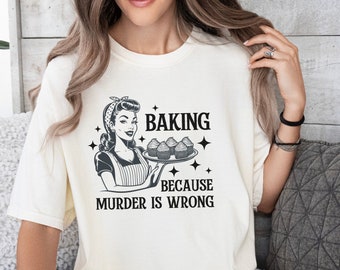 May include: A white t-shirt with a retro-style illustration of a woman holding a tray of cupcakes. The text on the shirt reads "Baking because murder is wrong."