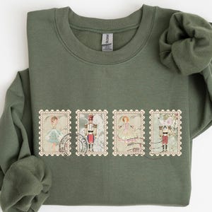 May include: An olive green sweatshirt featuring four postage stamp designs. Each stamp depicts a character from a ballet, including a ballerina, a toy soldier, and a mouse. The stamps have a vintage aesthetic with a decorative border.