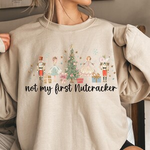 May include: Beige sweatshirt with a festive Nutcracker design. The graphic includes a Christmas tree, nutcrackers, and the text "not my first Nutcracker". A cozy, long-sleeved top for the holiday season.