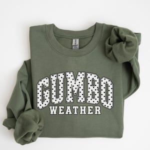 May include: A military green crewneck sweatshirt featuring the text GUMBO WEATHER in a collegiate font. The word GUMBO is filled with a white and black polka dot pattern, while the word WEATHER is in solid white letters with a black outline.