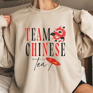 May include: Beige sweatshirt with the text "TEAM CHINESE Tea" in black and red. A red dragon graphic and a red and yellow umbrella are also featured on the front of the sweatshirt.