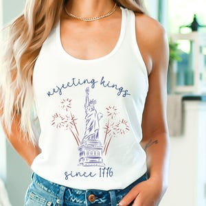 Pode incluir: Top branco sem mangas com um gráfico da Estátua da Liberdade, fogos de artifício e o texto "rejecting kings since 1776" em azul marinho e vermelho. A camisa é sem mangas.