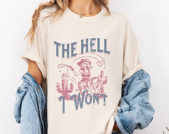 May include: A cream colored t-shirt with a vintage graphic of a woman in a cowboy hat holding a lasso. The text "THE HELL I WON'T" is printed on the shirt.