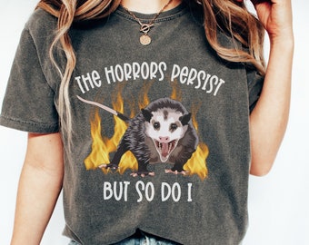 May include: A dark gray t-shirt with a graphic of a possum with flames behind it. The text on the shirt reads "The horrors persist but so do I."