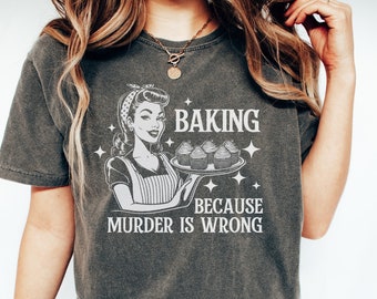 May include: A dark gray t-shirt with a retro-style illustration of a woman holding a tray of cupcakes. The text on the shirt reads "Baking because murder is wrong."