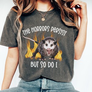K&ouml;nnte beinhalten: Ein dunkelgraues T-Shirt mit einem Aufdruck eines Opossums mit Flammen dahinter. Der Text auf dem T-Shirt lautet "The horrors persist but so do I."