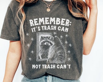 May include: A dark gray t-shirt with a white graphic of a raccoon looking in a mirror. The text "REMEMBER: IT'S TRASH CAN NOT TRASH CAN'T" is printed on the shirt.