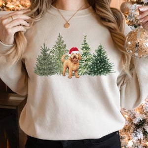 May include: A cream-colored sweatshirt with a Christmas-themed design featuring a golden doodle dog wearing a Santa hat standing in front of a row of decorated Christmas trees.
