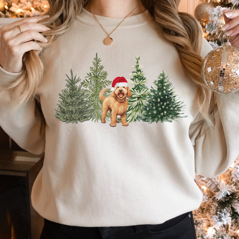 May include: A cream-colored sweatshirt with a Christmas-themed design featuring a golden doodle dog wearing a Santa hat standing in front of a row of decorated Christmas trees.