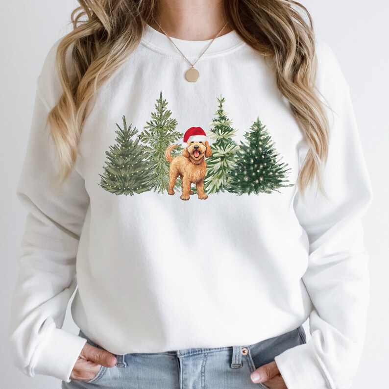 May include: White sweatshirt with a Christmas-themed design featuring a golden doodle dog wearing a Santa hat standing in front of a row of decorated Christmas trees.
