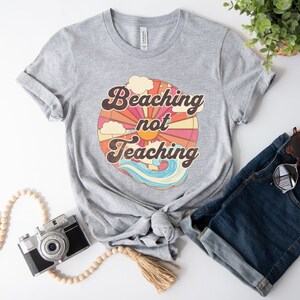 May include: Gray t-shirt with a retro-style graphic of a sun, clouds, and waves. The text "Beaching not Teaching" is printed on the shirt.