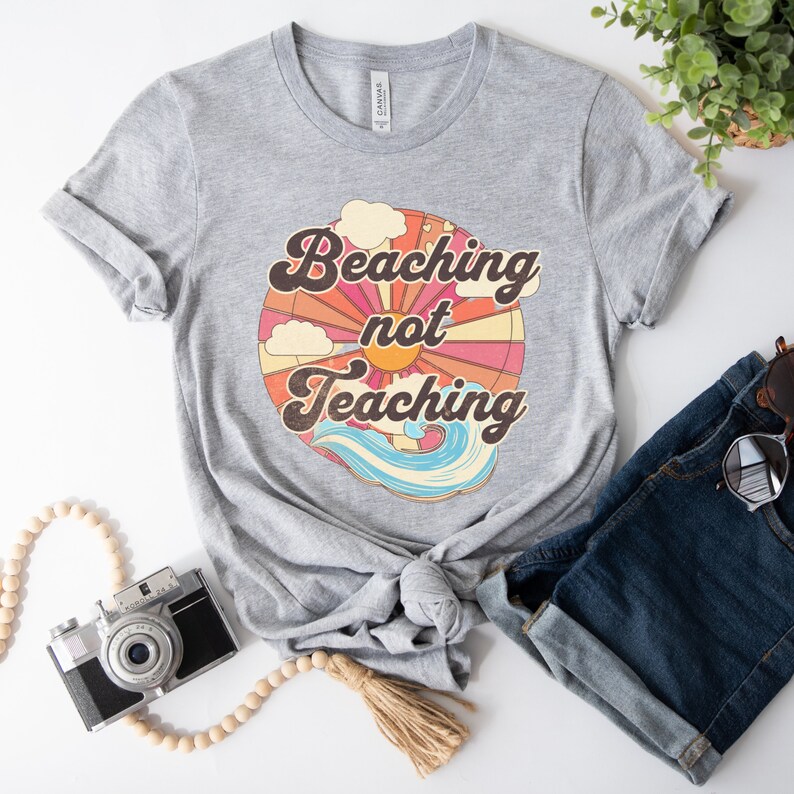 May include: Gray t-shirt with a retro-style graphic of a sun, clouds, and waves. The text "Beaching not Teaching" is printed on the shirt.