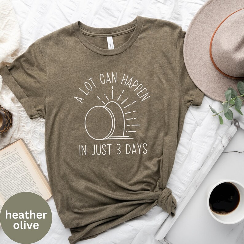 Pode incluir: Uma camiseta de cor oliva com um design gr&aacute;fico branco que diz "A lot can happen in just 3 days". O gr&aacute;fico apresenta uma imagem estilizada de um nascer do sol sobre um t&uacute;mulo.