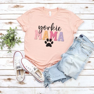 May include: A pink t-shirt with the text "yorkie MAMA" in rainbow colors and a black paw print. The shirt is laid out on a white wooden surface with a pair of white sneakers and blue jeans.