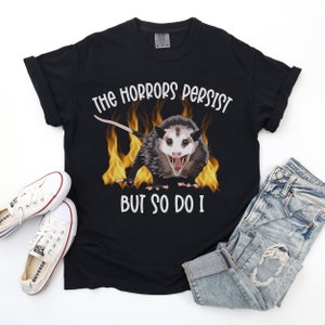 K&ouml;nnte beinhalten: Schwarzes T-Shirt mit einem Aufdruck eines Opossums in Flammen und dem Text "The horrors persist but so do I".