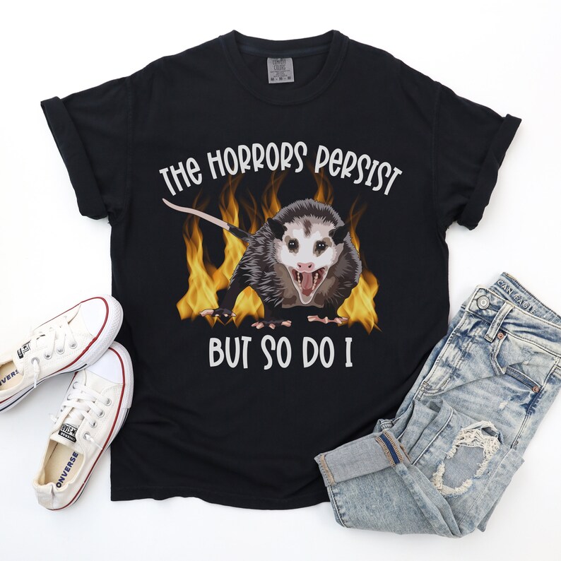 K&ouml;nnte beinhalten: Schwarzes T-Shirt mit einem Aufdruck eines Opossums in Flammen und dem Text "The horrors persist but so do I".