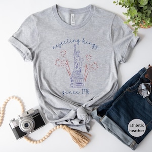 May include: Grey athletic heather t-shirt with a graphic of the Statue of Liberty, fireworks, and the text "rejecting kings since 1776" in blue and red. The shirt is knotted at the waist and paired with blue denim shorts.