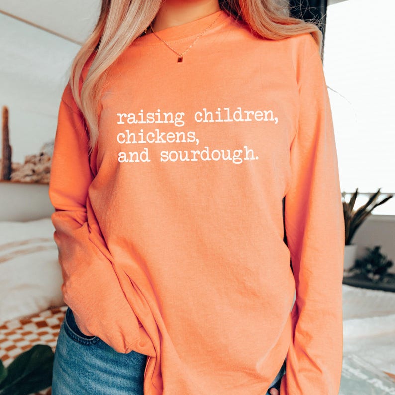 K&ouml;nnte beinhalten: Ein orangefarbenes Langarmshirt mit dem wei&szlig;en Text "raising children, chickens, and sourdough". Das Shirt hat eine einfarbige Farbe und einen Rundhalsausschnitt.