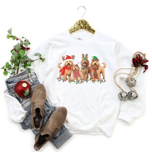 May include: White sweatshirt with a Christmas-themed design featuring four doodle dogs wearing festive hats and antlers. The dogs are standing in front of a string of Christmas lights.