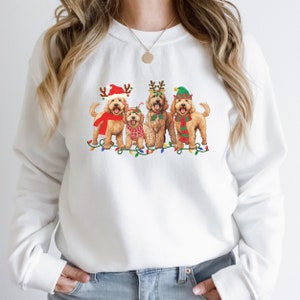 May include: White sweatshirt with a Christmas-themed design featuring four doodle dogs wearing festive hats and scarves. The dogs are standing in front of a string of Christmas lights.