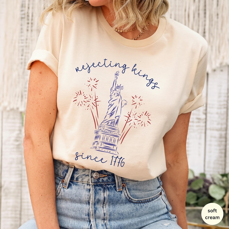 May include: A cream-coloured t-shirt with a navy blue illustration of the Statue of Liberty, fireworks, and the text "rejecting kings since 1776". The design is in navy blue and red.