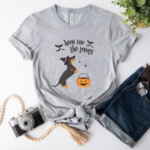 K&ouml;nnte beinhalten: Graues T-Shirt mit einem schwarzen und braunen Dackel, der einen Hexenhut tr&auml;gt und einen K&uuml;rbis-Eimer h&auml;lt. Der Text "here for the treats" ist auf dem Shirt aufgedruckt.
