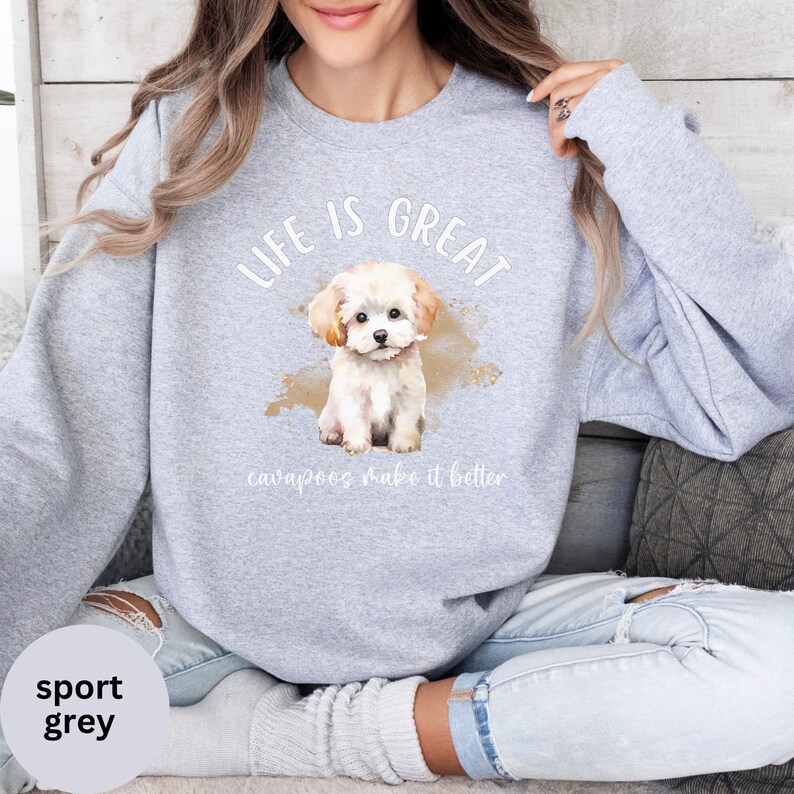 K&ouml;nnte beinhalten: Ein graues Sweatshirt mit dem Text "Life is Great" und einer Aquarell-Illustration eines wei&szlig;en Cavapoo-Welpen. Der Text "cavapoos make it better" befindet sich unter dem Welpen.