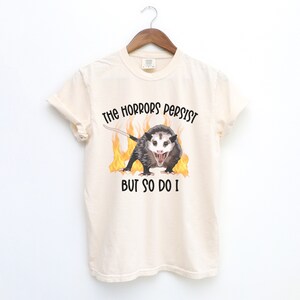 K&ouml;nnte beinhalten: Ein beiges T-Shirt mit einem Aufdruck eines Opossums in Flammen und dem Text "The horrors persist but so do I."