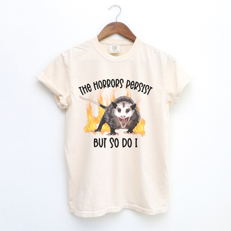 K&ouml;nnte beinhalten: Ein beiges T-Shirt mit einem Aufdruck eines Opossums in Flammen und dem Text "The horrors persist but so do I."