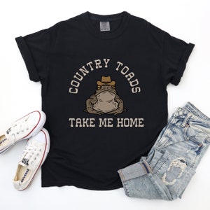 以下が含まれることがあります： カウボーイハットをかぶったカエルの漫画が描かれた黒いTシャツ。Tシャツには「Country Toads Take Me Home」という文字がプリントされています。