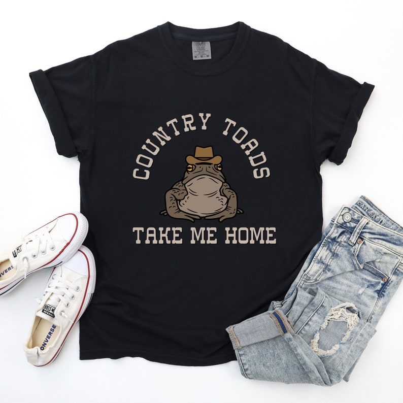 以下が含まれることがあります： カウボーイハットをかぶったカエルの漫画が描かれた黒いTシャツ。Tシャツには「Country Toads Take Me Home」という文字がプリントされています。