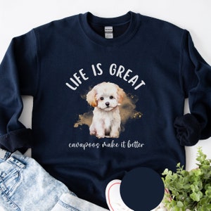 K&ouml;nnte beinhalten: Ein marineblauer Pullover mit einer wei&szlig;en Aquarell-Illustration eines Cavapoo-Welpen. Der Text "Life is great cavapoos make it better" ist auf dem Pullover aufgedruckt.