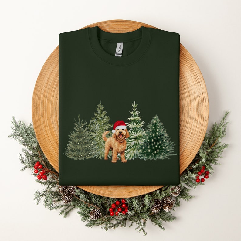 May include: A green t-shirt with a Christmas-themed design featuring a golden doodle dog wearing a Santa hat and standing in front of four decorated Christmas trees.