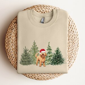 May include: A beige sweatshirt with a Christmas-themed design featuring a golden doodle dog wearing a Santa hat standing in front of a row of decorated Christmas trees.