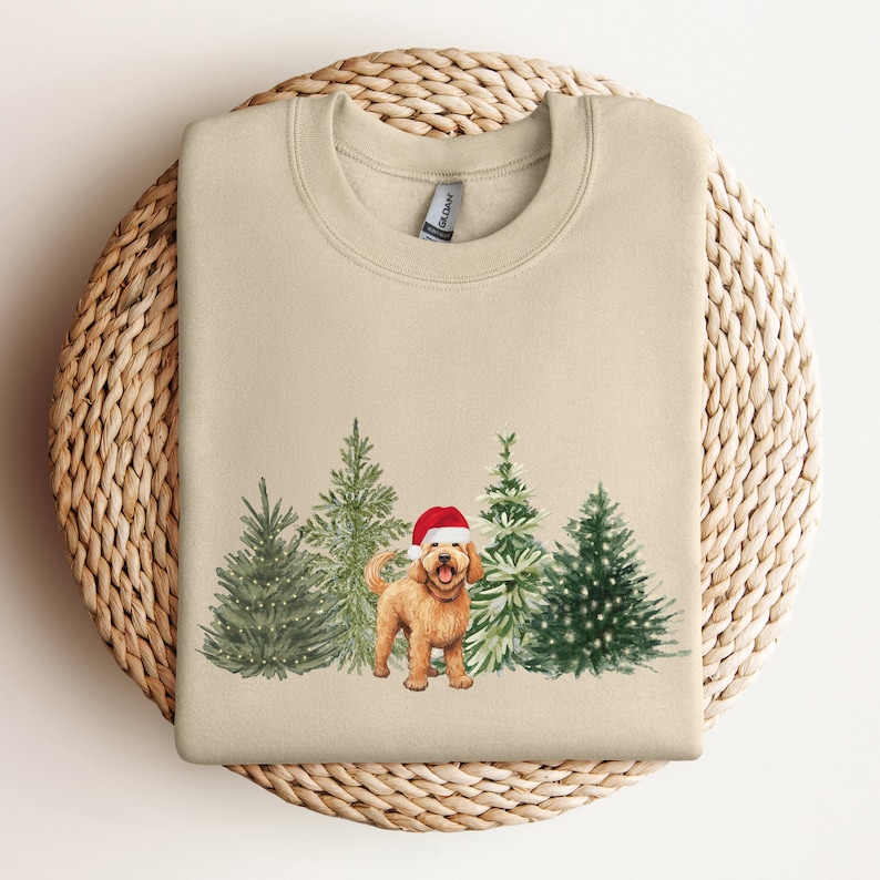May include: A beige sweatshirt with a Christmas-themed design featuring a golden doodle dog wearing a Santa hat standing in front of a row of decorated Christmas trees.
