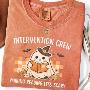 May include: A rust-colored t-shirt featuring a graphic of a ghost wearing a witch hat and reading an orange book. The text reads INTERVENTION CREW and MAKING READING LESS SCARY. The design includes small bats, flowers, and a checkered background.