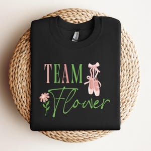 May include: Black sweatshirt with the words "TEAM Flower" in pink and green script. The design includes a pink flower and a pair of ballet shoes. The sweatshirt is folded on a woven, circular surface.