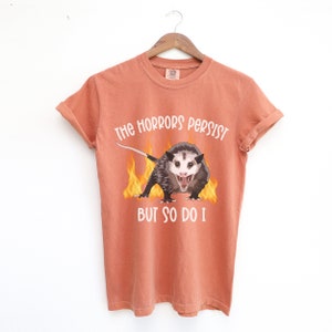 K&ouml;nnte beinhalten: Orangefarbenes T-Shirt mit einem Aufdruck eines Opossums mit Flammen dahinter. Der Text auf dem T-Shirt lautet "The horrors persist but so do I."
