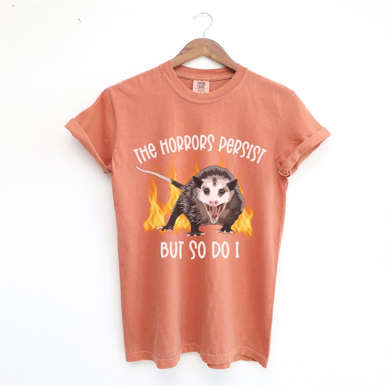 K&ouml;nnte beinhalten: Orangefarbenes T-Shirt mit einem Aufdruck eines Opossums mit Flammen dahinter. Der Text auf dem T-Shirt lautet "The horrors persist but so do I."