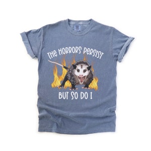 K&ouml;nnte beinhalten: Ein blaues T-Shirt mit einem Aufdruck eines Opossums in Flammen und dem Text "The horrors persist but so do I."