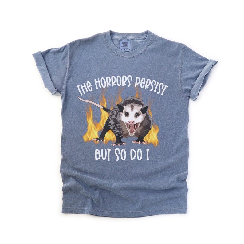K&ouml;nnte beinhalten: Ein blaues T-Shirt mit einem Aufdruck eines Opossums in Flammen und dem Text "The horrors persist but so do I."