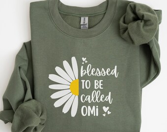May include: An olive green sweatshirt with a daisy graphic and the text "blessed to be called OMI." The daisy has white petals and a yellow center. The sweatshirt is folded with the sleeves bunched up.