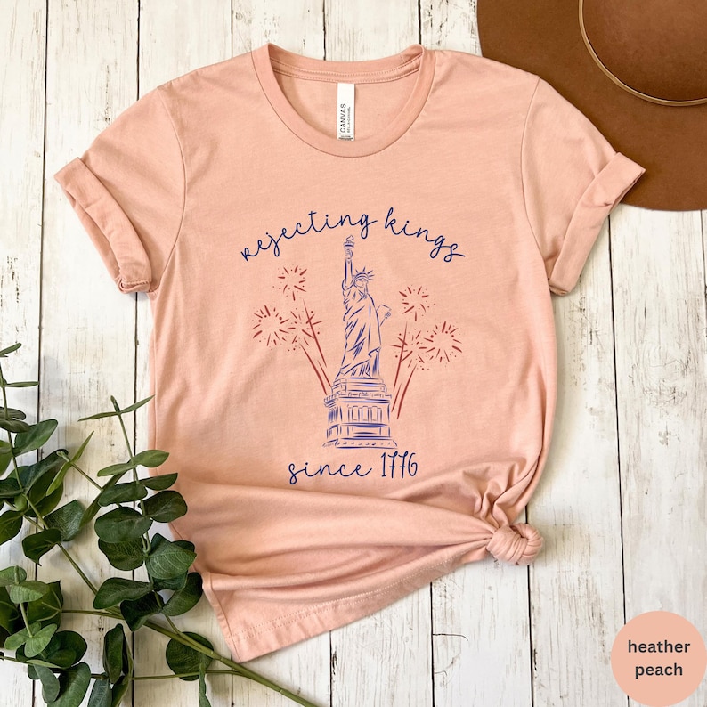 May include: Peach-coloured t-shirt featuring a navy blue graphic of the Statue of Liberty, fireworks, and the text "rejecting kings since 1776". The shirt is made of a soft material.