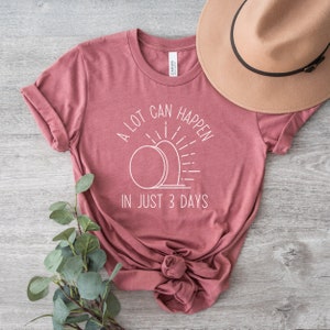 Pode incluir: Uma camiseta marrom-avermelhada com um design gr&aacute;fico branco que diz "Muita coisa pode acontecer em apenas 3 dias". O gr&aacute;fico apresenta uma imagem estilizada de um nascer do sol sobre um t&uacute;mulo.