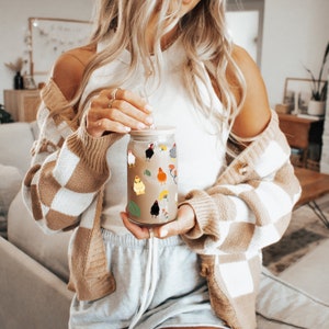 May include: A clear glass jar with a straw and a brown lid, decorated with colorful chicken stickers. The jar is being held by a person wearing a brown and white cardigan.