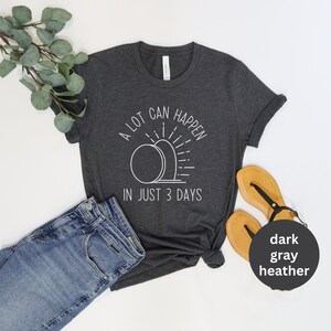 Pode incluir: Uma camiseta cinza heather escura com um gr&aacute;fico branco de um nascer do sol sobre duas l&aacute;pides. O texto na camiseta diz "A lot can happen in just 3 days."