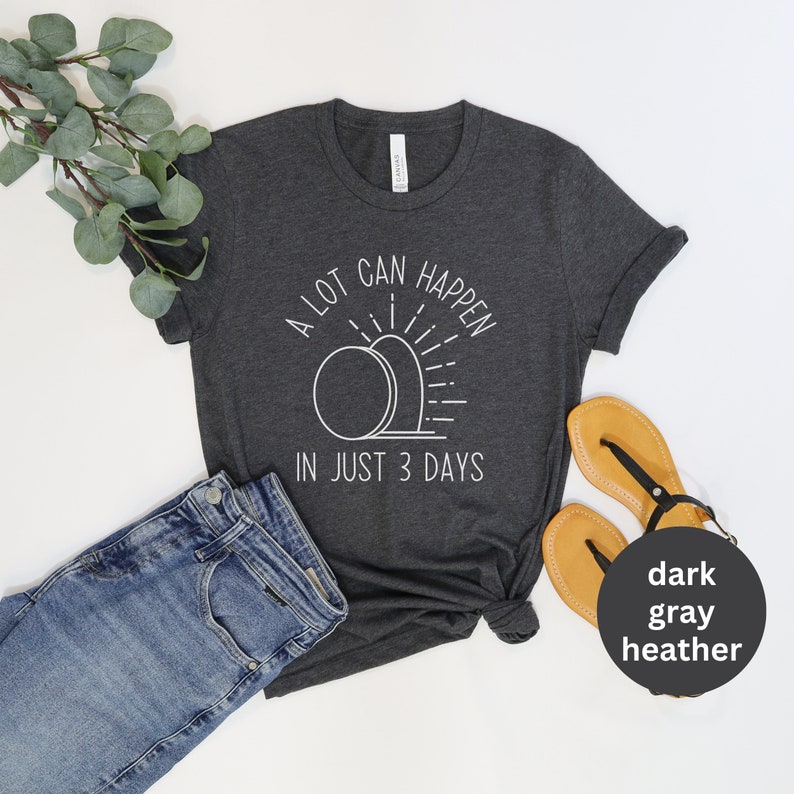 Pode incluir: Uma camiseta cinza heather escura com um gr&aacute;fico branco de um nascer do sol sobre duas l&aacute;pides. O texto na camiseta diz "A lot can happen in just 3 days."