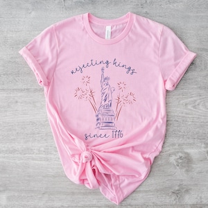 May include: A pale pink t-shirt with a graphic of the Statue of Liberty, fireworks, and the text "rejecting kings since 1776" in blue script. The shirt is made of soft material and has rolled-up sleeves.