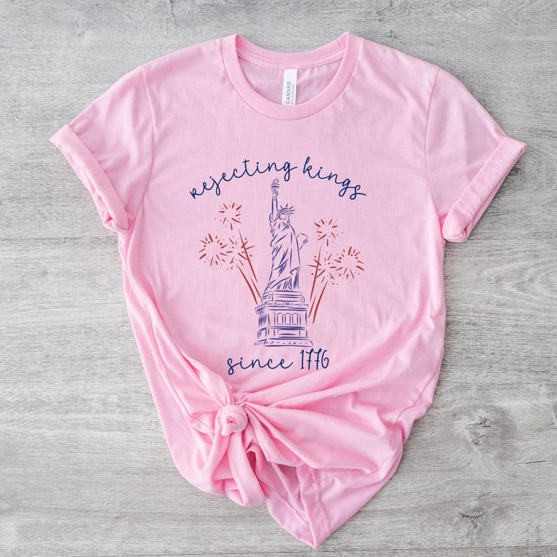 May include: A pale pink t-shirt with a graphic of the Statue of Liberty, fireworks, and the text "rejecting kings since 1776" in blue script. The shirt is made of soft material and has rolled-up sleeves.