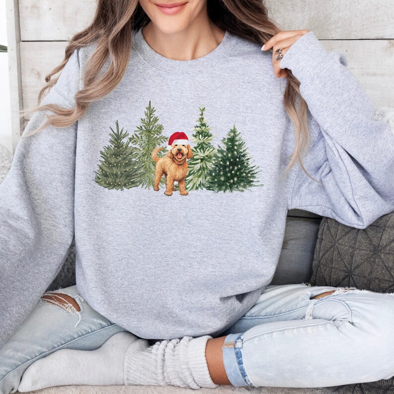 May include: A gray sweatshirt with a Christmas-themed design featuring a golden doodle dog wearing a Santa hat standing in front of a row of decorated Christmas trees.